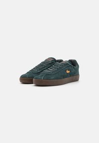 Green suede sneakers with a brown rubber sole, textured upper, and lace closure. Features a small orange logo on the side.