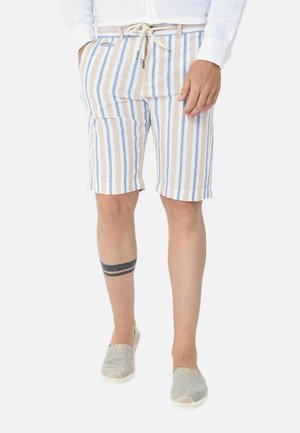 Uomo che indossa pantaloncini beige, bianchi e blu a righe verticali con coulisse in vita, scarpe grigie slip-on e un tatuaggio a doppia fascia nera sul polpaccio sinistro.
