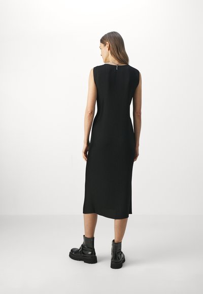 Robe noire sans manches avec texture côtelée verticale, silhouette ajustée, longueur au genou et un petit zip à l'arrière. Associée à des bottines noires épaisses.