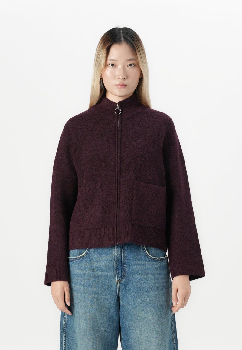 Kvinne iført en mørk lilla zip-up genser med lommer og løse blå jeans, stående mot en helt hvit bakgrunn.