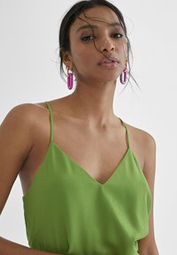 Groene satijnen top met dunne banden en een V-hals. Opvallende paarse hoepeloorbellen voegen een vleugje kleur toe. Soepele textuur, minimalistisch ontwerp.