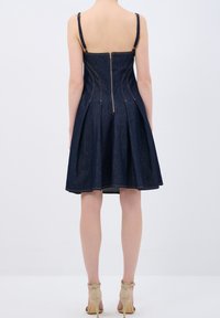 Jeanskleid mit tailliertem Oberteil, plissiertem Rock, Reißverschluss am Rücken und verstellbaren Trägern. Dunkelblaue Farbe mit goldenen Nahtdetails.