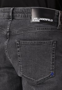 Jeans din denim gri închis, având o etichetă neagră cu inscripția "KARL LAGERFELD JEANS." Culoare albastră notabilă pe cusătura din spatele buzunarului.