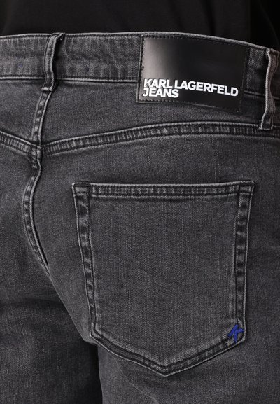 Jean en denim gris foncé avec une étiquette noire portant "KARL LAGERFELD JEANS". Couture bleue distinctive sur la poche arrière.