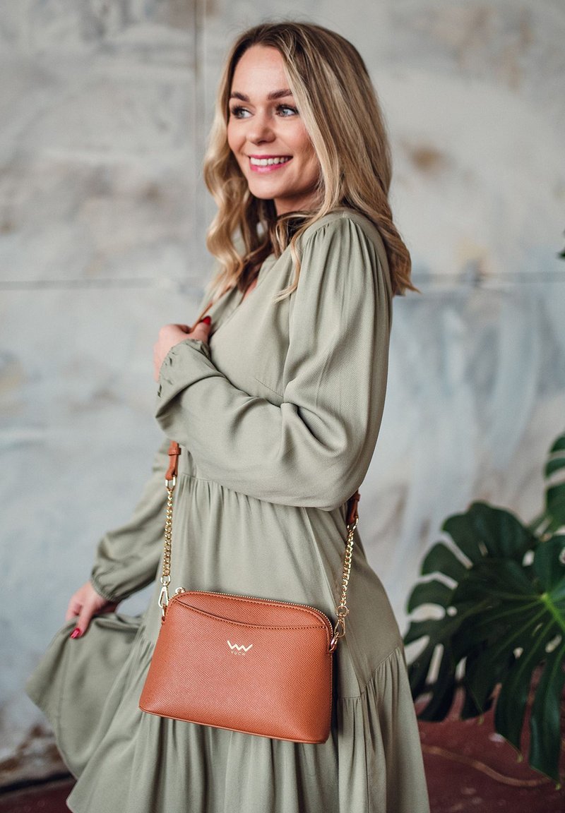 Braune, strukturierte Umhängetasche mit einem Reißverschluss, goldenen Beschlägen und einem Kettenriemen, getragen über einem grünen Kleid vor einem gedämpften Hintergrund.