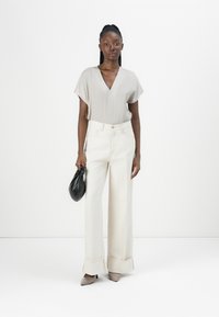 Crème wijde broek met een cropped zoom, gecombineerd met een lichtgrijs V-hals top. Het model houdt een zwarte handtas vast en draagt beige hakken.