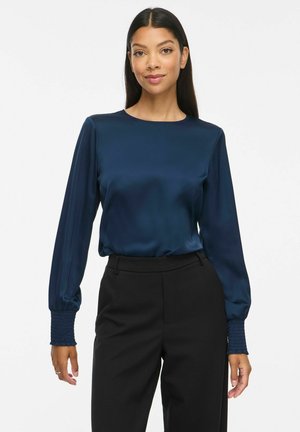 Femme aux longs cheveux foncés porte un chemisier en satin bleu marine à manches longues et un pantalon noir taille haute sur un fond blanc uni.