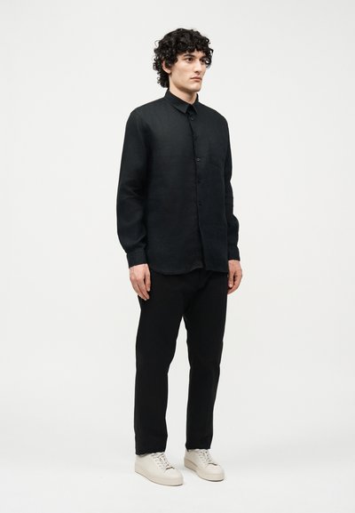 Άνδρας με κατσαρά μαλλιά στέκεται στραμμένος προς τα εμπρός, φορώντας μαύρο πουκάμισο με κουμπιά, μαύρο παντελόνι και λευκά αθλητικά παπούτσια μπροστά σε απλό λευκό φόντο.