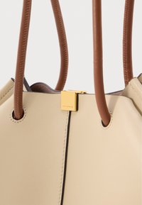 Μπεζ δερμάτινη τσάντα χειρός με καφέ στρογγυλές λαβές και μικρό χρυσό τετράγωνο κούμπωμα με ανάγλυφη την επιγραφή "COCCINELLE" στο μπροστινό μέρος.