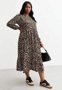 Maxi-jurk met dierenprint in beige en bruin, met lange mouwen, geplooide taille en gelaagde rok; gedragen met zwarte sneakers en een zwarte tas.
