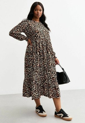 Maxi-jurk met dierenprint in beige en bruin, met lange mouwen, geplooide taille en gelaagde rok; gedragen met zwarte sneakers en een zwarte tas.