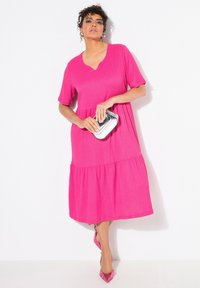 Fuksia mekko, jossa on lyhyet hihat, V-pääntie ja kerrostettu helma. Tyylitelty hopeisella clutchilla ja vaaleanpunaisilla kärkitossuilla. Sileä pinta.