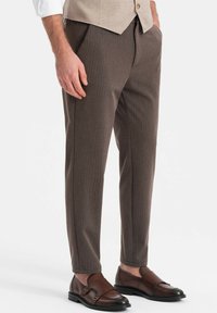 Pantalones marrones con corte a medida y un sutil patrón de rayas verticales. Presentan bolsillos laterales, un corte relajado y dobladillos ajustados. Combinados con zapatos de cuero marrones.