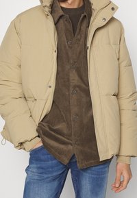 Beige puffer jacket with side pockets and a high collar, worn over a brown corduroy shirt. The outfit is paired with blue denim jeans.
