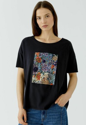 Femme aux longs cheveux bruns portant un t-shirt noir avec un patch floral coloré et un jean bleu, debout avec une main dans la poche.