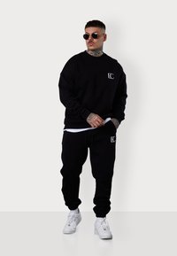 Schwarzes Sweatshirt- und Jogginganzug-Set aus Baumwolle. Mit Logo-Details auf der Brust und dem Oberschenkel, kombiniert mit weißen Sneakers und runden Sonnenbrillen.