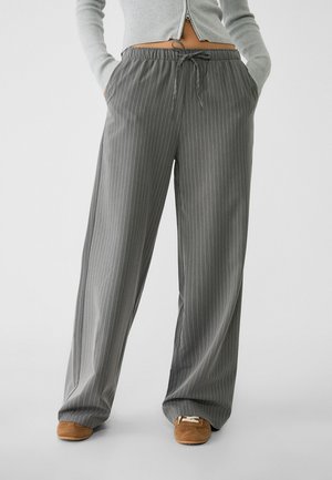 Personne portant un pantalon gris à rayures larges avec taille à cordon et chaussures marron, mains dans les poches, debout devant un fond uni.