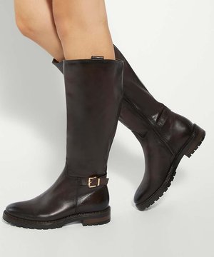 Bottes hautes en cuir brun foncé brillant avec détail de boucle latérale et semelle en caoutchouc épaisse portées sur des jambes croisées sur un fond clair.