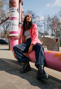Blazer rosa sopra una canotta grigia, pantaloni a quadri blu scuro e stivali platform neri. Seduta su un tubo rosa in un contesto all'aperto.