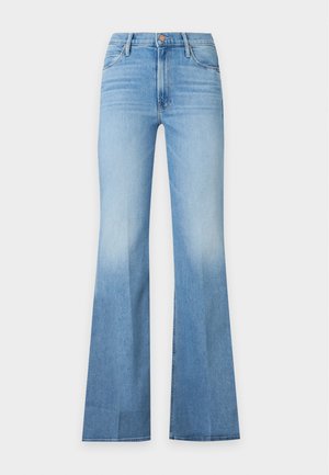 Femme debout, main dans la poche, portant un col roulé blanc, un jean évasé bleu clair et des chaussures noires pointues sur un fond uni.