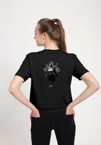 Žena s hnědým ohonem má na sobě černé tričko Diablo IV s designem zakryté temné postavy na zádech, stojí na bílém pozadí.