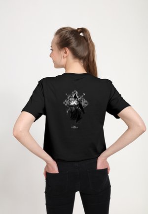 Žena s hnědým ohonem má na sobě černé tričko Diablo IV s designem zakryté temné postavy na zádech, stojí na bílém pozadí.