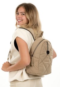 Jauna sieviete smaidot pār plecu, valkājot baltu bezroku vilnas džemperi un nēsājot bēša krāsas rūtainu mugursomu ar rāvējslēdzēju un ķēdes detaļu.