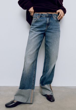 Helle blaue Denim weit geschnittene Jeans mit ausgefransten Säumen, featuring kontrastierenden Nähten und einem lässigen Schnitt. Kombiniert mit dunklen flachen Schuhen.