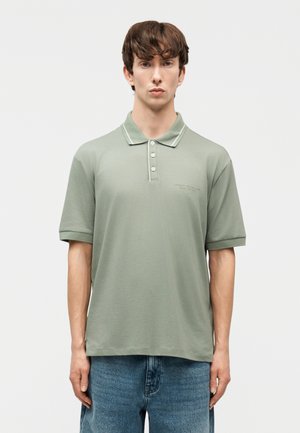 Jeune homme aux cheveux bruns portant un polo à manches courtes vert clair et un jean bleu, debout devant un fond blanc uni.