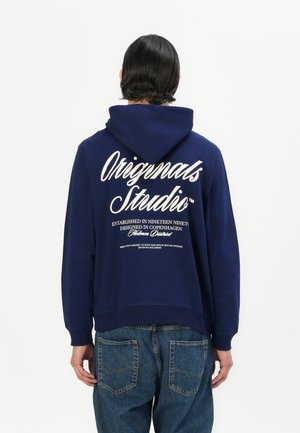Hanorac albastru închis cu un imprimeu mare alb "Originals Studio" pe spate. Dispune de un glugă cu șnur și tivuri și manșete dintr-un material ribat. Purtați cu blugi de denim.