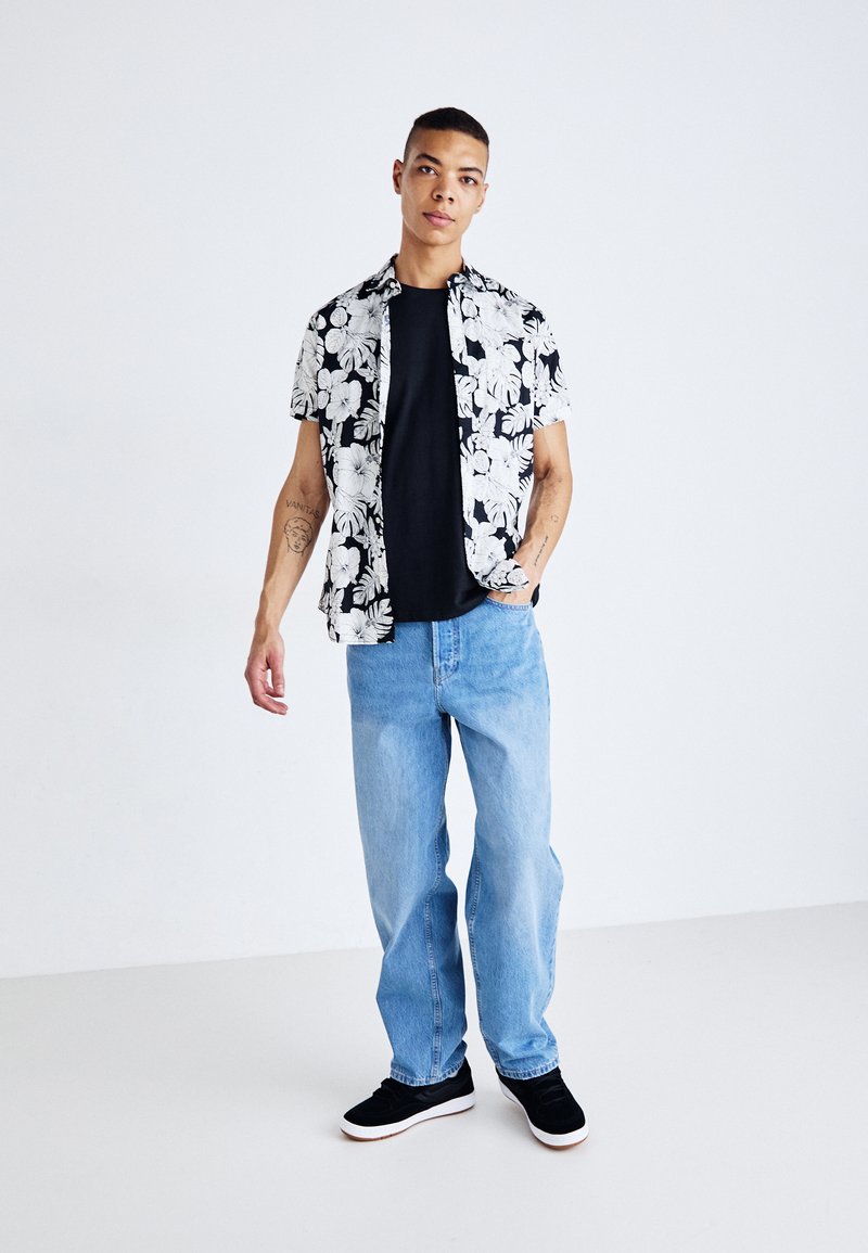 Korte mouwen zwart-wit bloemenoverhemd, zwart T-shirt, lichtblauwe losse jeans en zwarte sneakers met witte zolen.