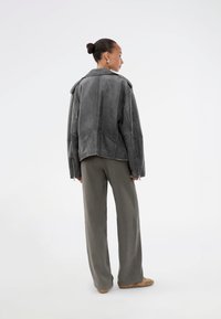 Veste en cuir grise au design structuré, avec des coutures et des zips proéminents, associée à un large pantalon vert et des chaussures beiges.