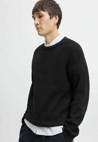 Zwarte geribbelde gebreide trui met een ronde hals en lange mouwen. Draagt over een witte gestreepte blouse met kraag, wat een gelaagde look geeft.