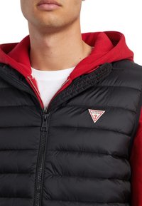 Chaleco acolchado negro con cierre de cremallera y sudadera con capucha roja debajo. Cuenta con un pequeño logo triangular rojo y blanco en el pecho.