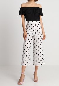 Haut noir à épaules dénudées avec bordure à volants, associé à un pantalon blanc à jambes larges orné de gros pois noirs. Talons roses à bride cheville.