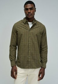 Zielona koszula flanelowa w kratę z kołnierzykiem zapinanym na guziki, długimi rękawami i zapięciem na guziki z przodu. Nosi się ją na białej koszuli, w połączeniu z jasnymi spodniami.