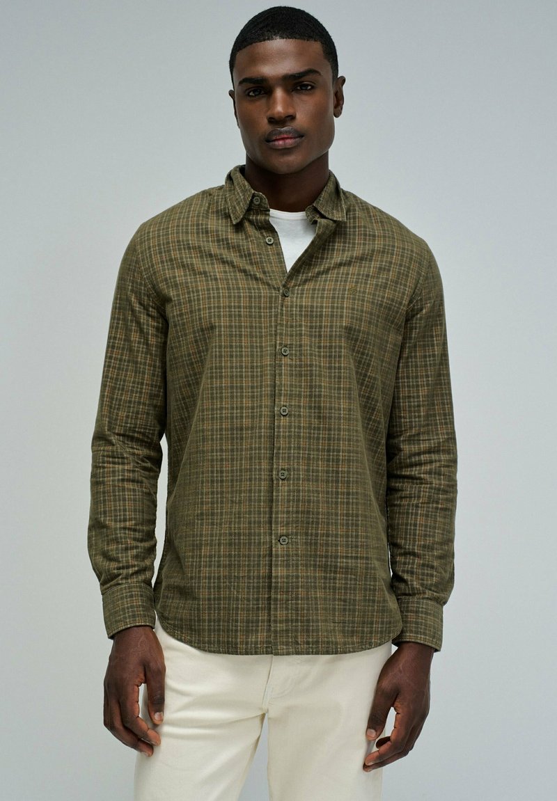 Zielona koszula flanelowa w kratę z kołnierzykiem zapinanym na guziki, długimi rękawami i zapięciem na guziki z przodu. Nosi się ją na białej koszuli, w połączeniu z jasnymi spodniami.