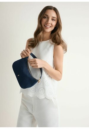 Młoda kobieta w białym stroju uśmiecha się, trzymając granatową torebkę na suwak na neutralnym tle.