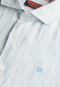 Lichtblauw gestreept overhemd van een zachte stof, met een knoopsluiting bij de kraag en een klein geborduurd logo in het blauw op de linkerkant van de borst.