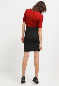 Haut rouge à manches courtes côtelées associé à une jupe noire ajustée à motif en chevrons. Des escarpins noirs à talons aiguilles complètent la tenue.