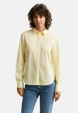 Frau mit lockigem Haar, die ein hellgelbes Hemd mit Knöpfen und blaue Jeans trägt, steht vor einem schlichten hellen Hintergrund.