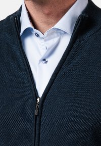 Námořnicky modrý svetr na zip s texturovaným pleteným vzorem, vybavený stříbrným zipem a vrstvený přes světle modrou košili s límečkem.