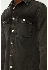 Camicia in denim nero con chiusura a bottoni, due tasche sul petto, ferrature argento, cuciture a contrasto e colletto classico.