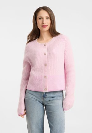 Frau trägt eine weiche rosa Knopf-Cardigan mit langen Ärmeln und hellblauen Jeans, stehend vor einem schlichten weißen Hintergrund.