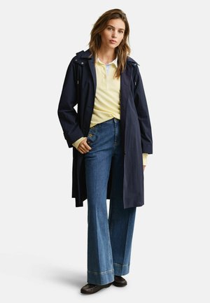 Woman wearing navy raincoat over light yellow polo shirt and high-waisted wide-leg jeans with button details, standing against white background.