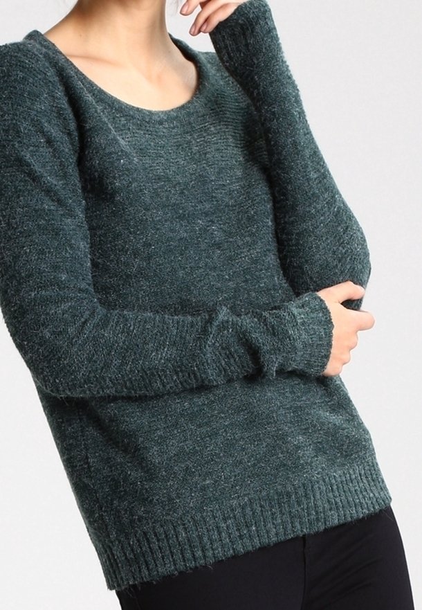Personne portant un pull en laine vert foncé à manches longues avec des poignets et un ourlet côtelés, les bras croisés, cadrée au niveau du cou et des hanches.