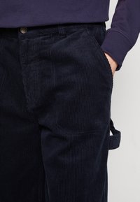 Pantalones de pana azul marino con textura de ribete vertical, bolsillo lateral y detalle de trabilla, que presentan un corte clásico y sutiles acentos de costura.