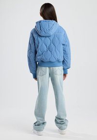 Blue quilted jacket with a hood, featuring a textured pattern and ribbed cuffs, paired with light-wash straight-leg jeans and white sneakers.