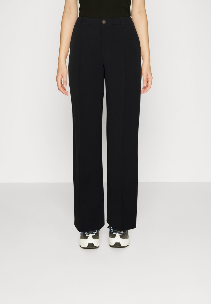 Femme portant un pantalon noir à jambes larges et des baskets blanches, debout devant un fond clair uni.