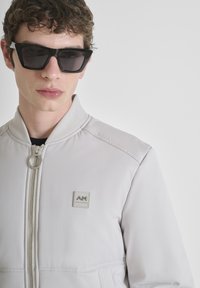 Jongeman in zwarte rechthoekige zonnebril en een lichtgrijze zip-up jas met een klein logo op de borst, neutrale achtergrond.